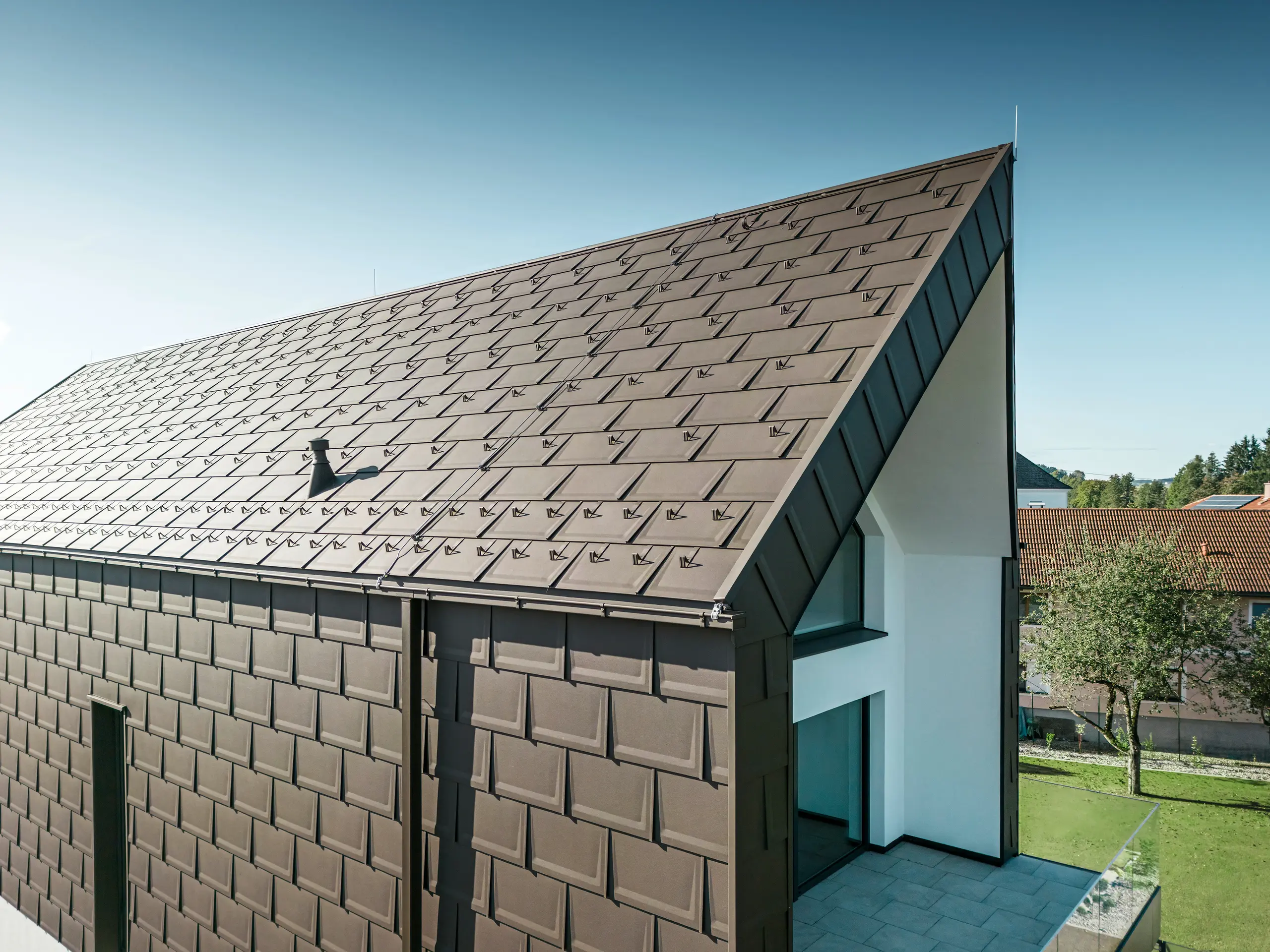 Eine perspektivische Ansicht von Dach und Fassade eines modernen Einfamilienhauses in Neukirchen, Österreich. Die PREFA R.16 Dachplatte im Braunton P.10 bietet eine ästhetische und funktionale Dacheindeckung, die sich perfekt in die architektonische Gestaltung des Hauses einfügt. Der scharfe First des Daches und die präzise Anordnung der Dachplatten zeigen die Qualität von PREFA. Die kleinformatigen Aluminiumprodukte fügen sich perfekt in die weiß verputzte Fassade und die umgebende grüne Landschaft ein.