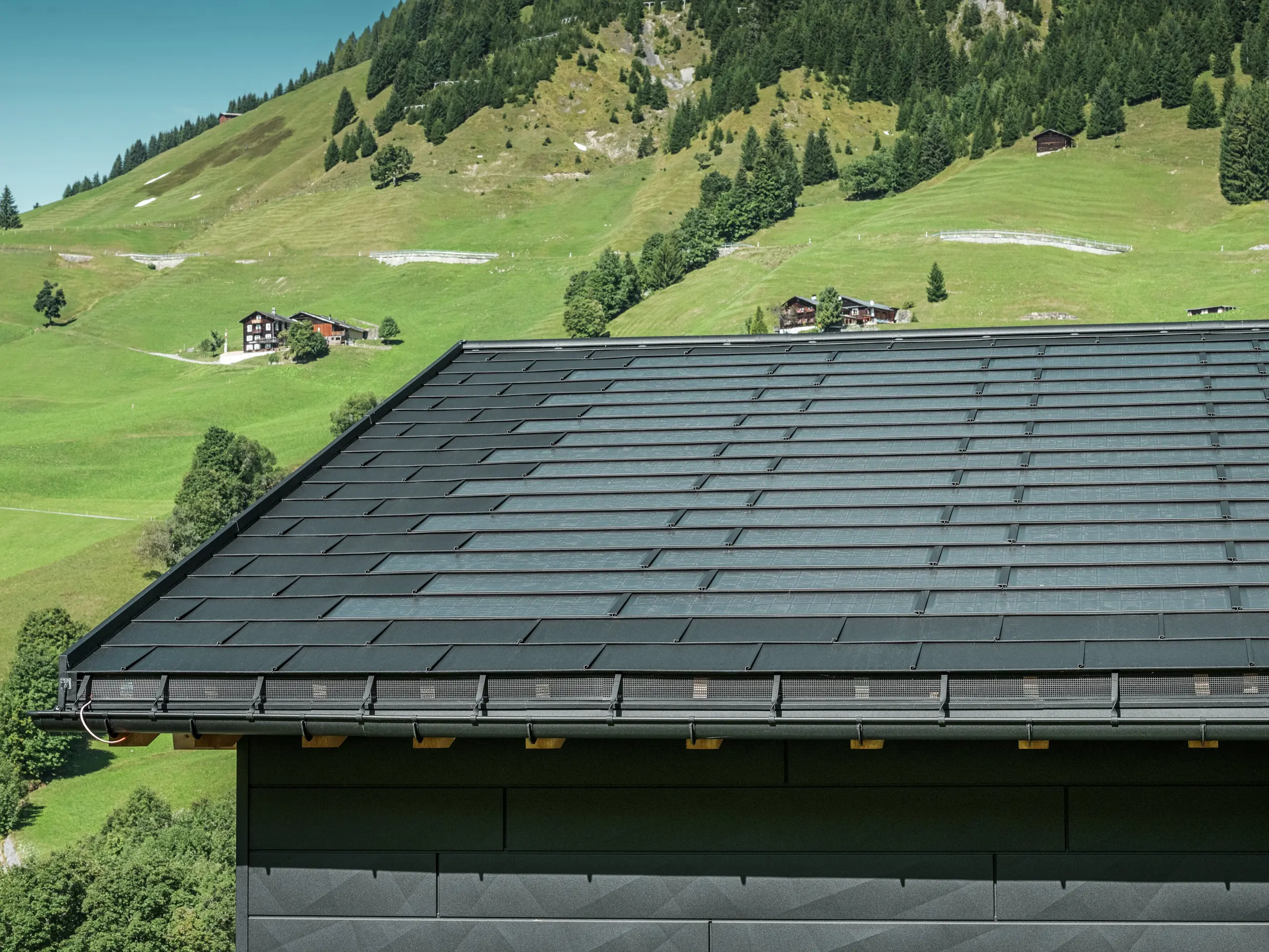 Nahaufnahme der Traufe eines modernen Einfamilienhauses in der alpinen Umgebung von Ascharina in der Schweiz. Das Dach wurde mit der innovativen PREFA Solardachplatte und der traditionellen Dachplatte R.16, beide in P.10 Schwarz, eingedeckt. Für die zuverlässige Dachentwässerung sorgen die PREFA Hängerinne und das PREFA Ablaufrohr. Am unteren Bildrand sind die unverwechselbaren PREFA Siding.X Fassadenpaneele zu sehen. Die klare Linienführung der Aluminiumprodukte unterstreicht das moderne Design inmitten einer malerischen Berglandschaft.