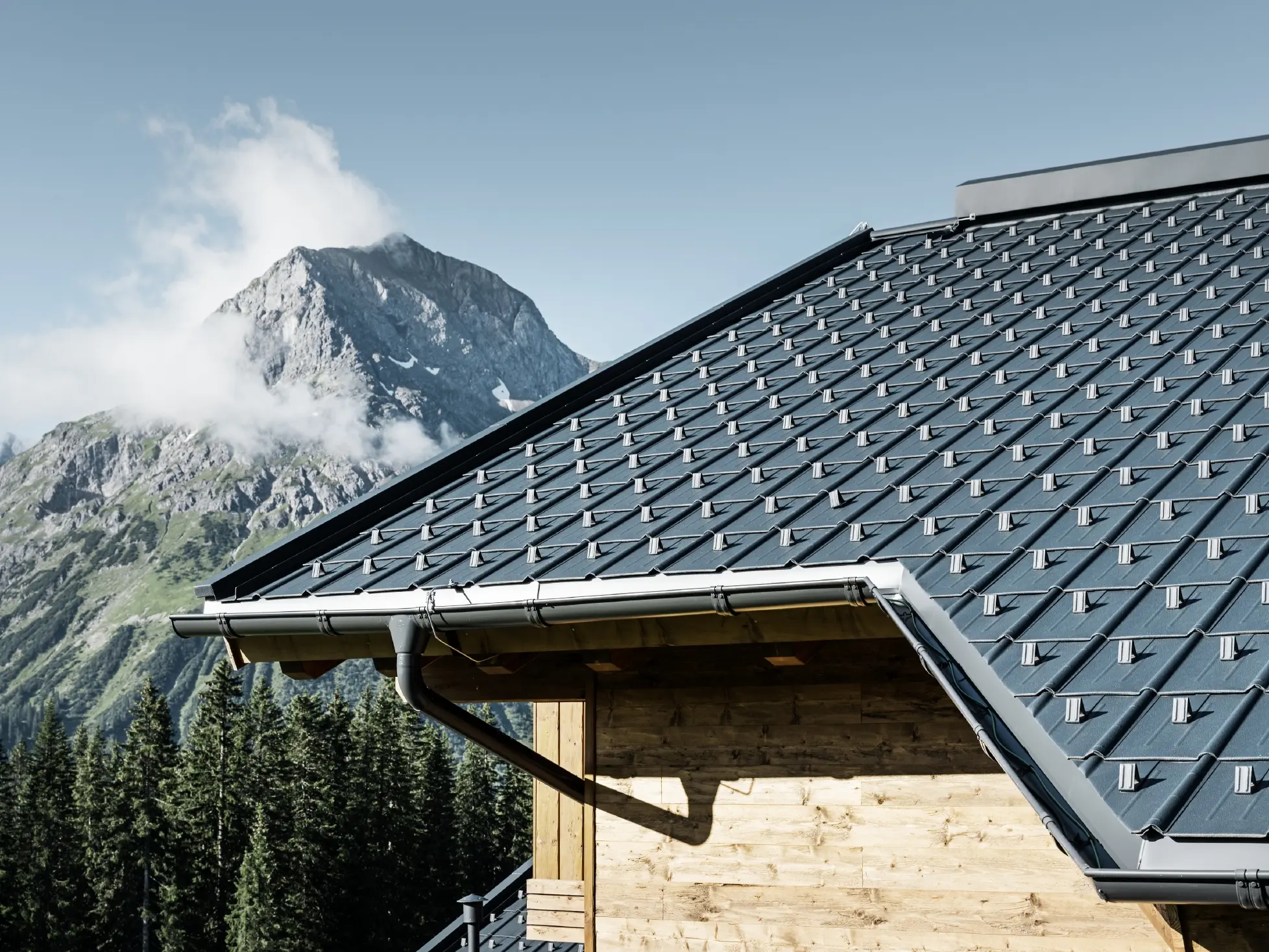 PREFA krovna ploča, primjena u alpskom okruženju – krovna ploča u boji antracit postavljena na kolibu u naselju Lech am Arlberg, s planinama u pozadini; PREFA krovni oluk u boji antracit; drvena fasada