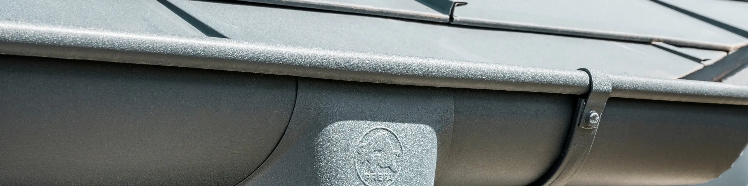 Krupni plan krovne konstrukcije kuće s PREFA krovnom pločom R.16 u antracit boji, kao i detaljan prikaz koloristički usklađenog PREFA krovnog oluka i odvodne cijevi od aluminijuma, pri sunčanom vremenu.