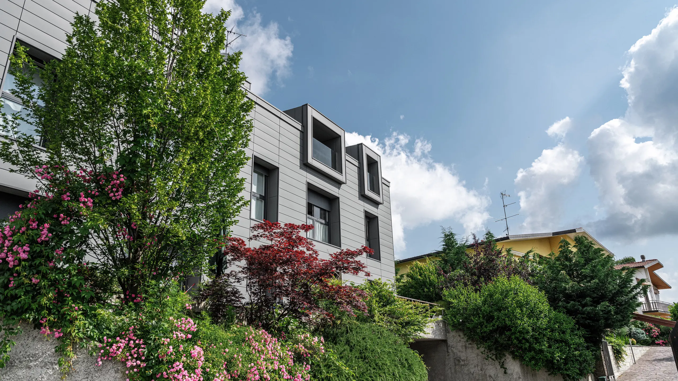 Moderne Wohnanlage in Faloppio, Italien, mit stilvoller Fassade aus PREFA Sidings. Die auskragenden Fenster bieten einen einzigartigen architektonischen Blickfang, während die umliegenden Pflanzen und Blumen die natürliche Schönheit des Objektes unterstreichen. Die langlebige Aluminiumverkleidung von PREFA sorgt für eine ästhetische und funktionelle Lösung bei jeder Witterung.